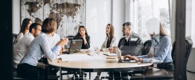 A group of people in business attire sit around a conference table in a modern office, discussing work with laptops and documents. A world map hangs on the wall in the background.