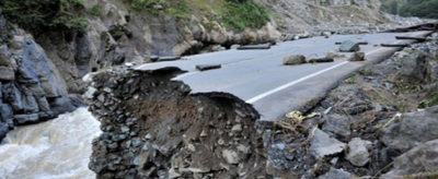 A section of paved road is collapsed into a river below, with jagged edges and loose debris visible, surrounded by rocky cliffs and greenery.