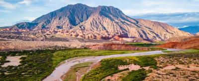 A winding river flows through a green valley with a large, rocky mountain in the background under a partly cloudy sky.