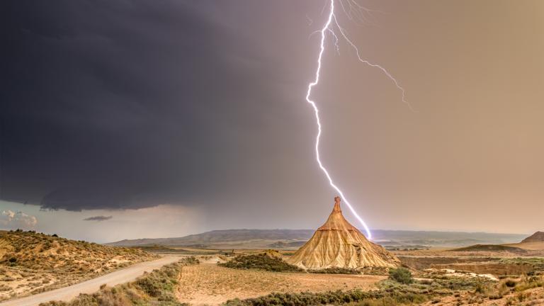 A bolt of lightning strikes a conical rock formation in a dry, open landscape under a dramatic, cloudy sky.