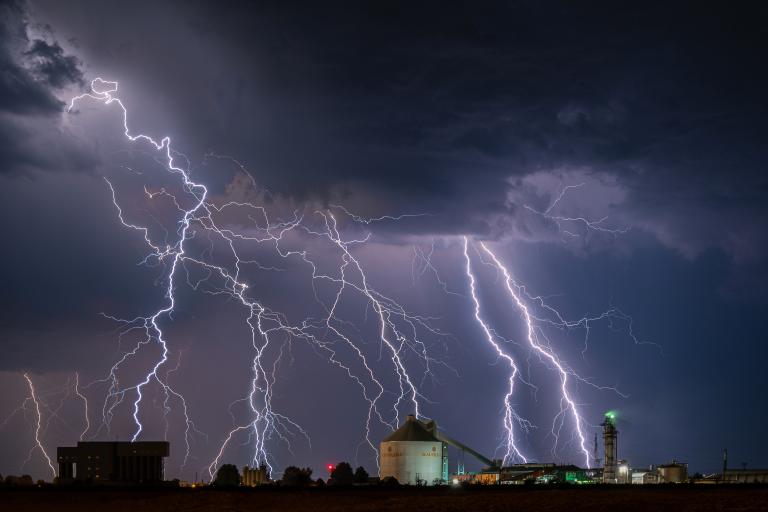 Multiple lightning bolts strike the ground near industrial buildings under a dark, cloudy night sky.