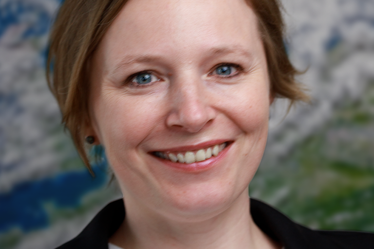 A woman with short blonde hair, wearing a black blazer and white shirt, smiles at the camera. A blurred image of Earth is in the background.