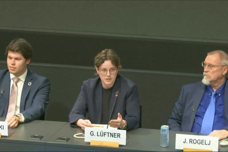 Three people sit at a panel table, with microphones and nameplates in front of them. The person in the center is speaking while the others listen.