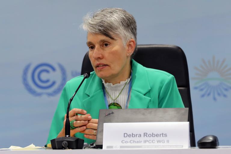 A woman in a green jacket speaks at a conference table with a microphone and nameplate, with conference logos visible in the background.
