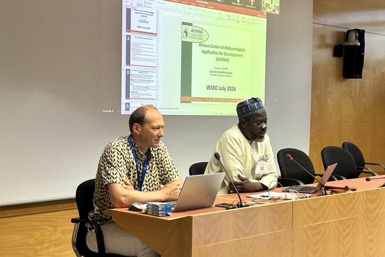 Two men sit at a conference table presenting in front of a screen displaying a slide titled "African Centre of Meteorological Applications for Development.