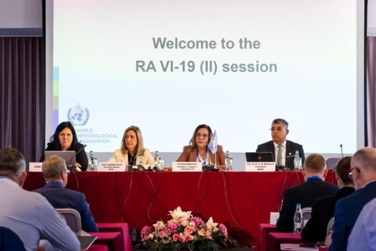 Four speakers sit at a red-covered table in front of a projection screen that reads “Welcome to the RA VI-19 (II) session.”