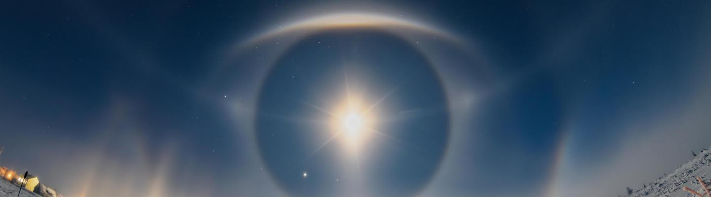 A snowy road under a night sky with a bright moon surrounded by halos and light pillars, creating concentric circular patterns.