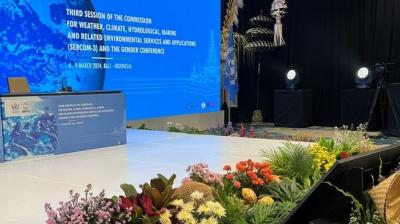 A conference stage with a large blue screen displaying information about the Third Session of the Commission for weather and climate in Bali, decorated with colorful flowers in the foreground.