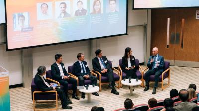 A panel of six people in business attire sit on stage discussing during a conference, with an audience watching and a presentation screen displayed above them.