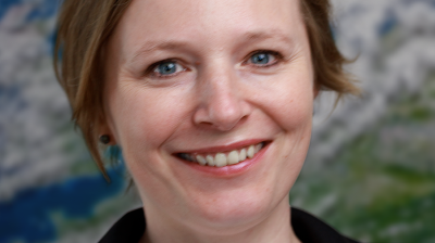 A woman with short blonde hair, wearing a black blazer and white shirt, smiles at the camera. A blurred image of Earth is in the background.