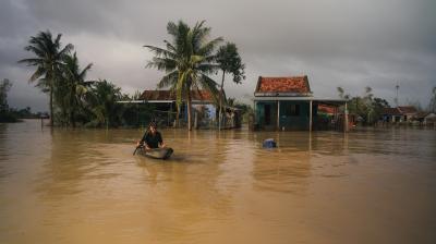 A person paddles a small boat through floodwaters in front of partially submerged houses and palm trees under a cloudy sky.