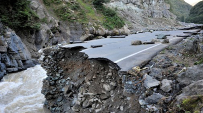 A section of paved road is collapsed into a river below, with jagged edges and loose debris visible, surrounded by rocky cliffs and greenery.