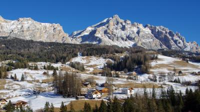 Snow-dusted mountains and scattered houses on a hillside under a clear blue sky, with patches of trees and grass visible.