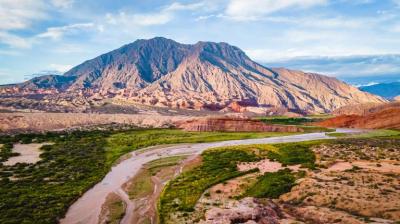 A winding river flows through a green valley with a large, rocky mountain in the background under a partly cloudy sky.