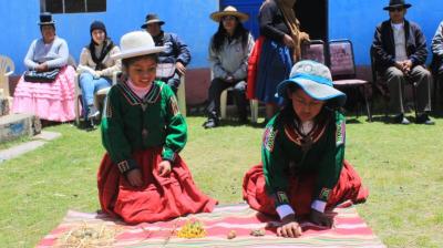 Two girls in traditional Andean clothing play a game with small objects on a woven mat outdoors, while people sit and watch in the background.