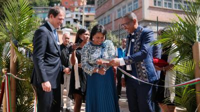 Three people stand smiling at an outdoor ribbon cutting ceremony, surrounded by others and greenery, with buildings visible in the background.