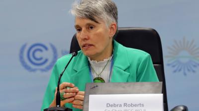 A woman in a green jacket speaks at a conference table with a microphone and nameplate, with conference logos visible in the background.