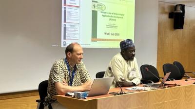 Two men sit at a conference table presenting in front of a screen displaying a slide titled "African Centre of Meteorological Applications for Development.