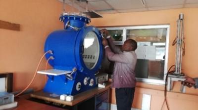 A person adjusts controls on a large blue scientific instrument in a laboratory with light orange walls and various equipment.