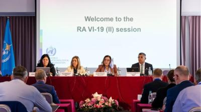 Four speakers sit at a red-covered table in front of a projection screen that reads “Welcome to the RA VI-19 (II) session.”