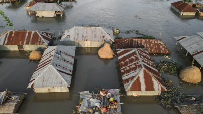 Aerial view of houses partially submerged in floodwater, with people on a makeshift raft in the center.