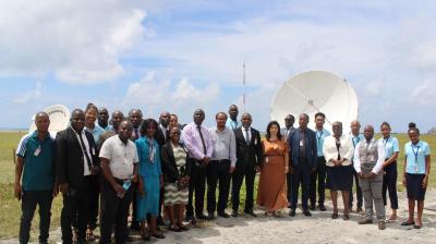 A group of people poses outdoors in front of satellite dishes on a grassy area, dressed in business and casual attire under a partly cloudy sky.