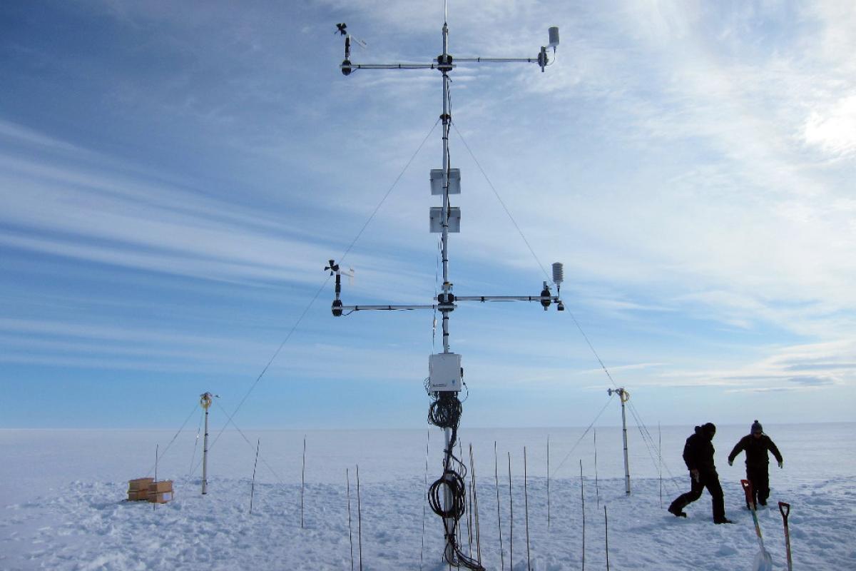 Two scientists working on a complex weather station apparatus on a snow-covered plain under a clear blue sky.