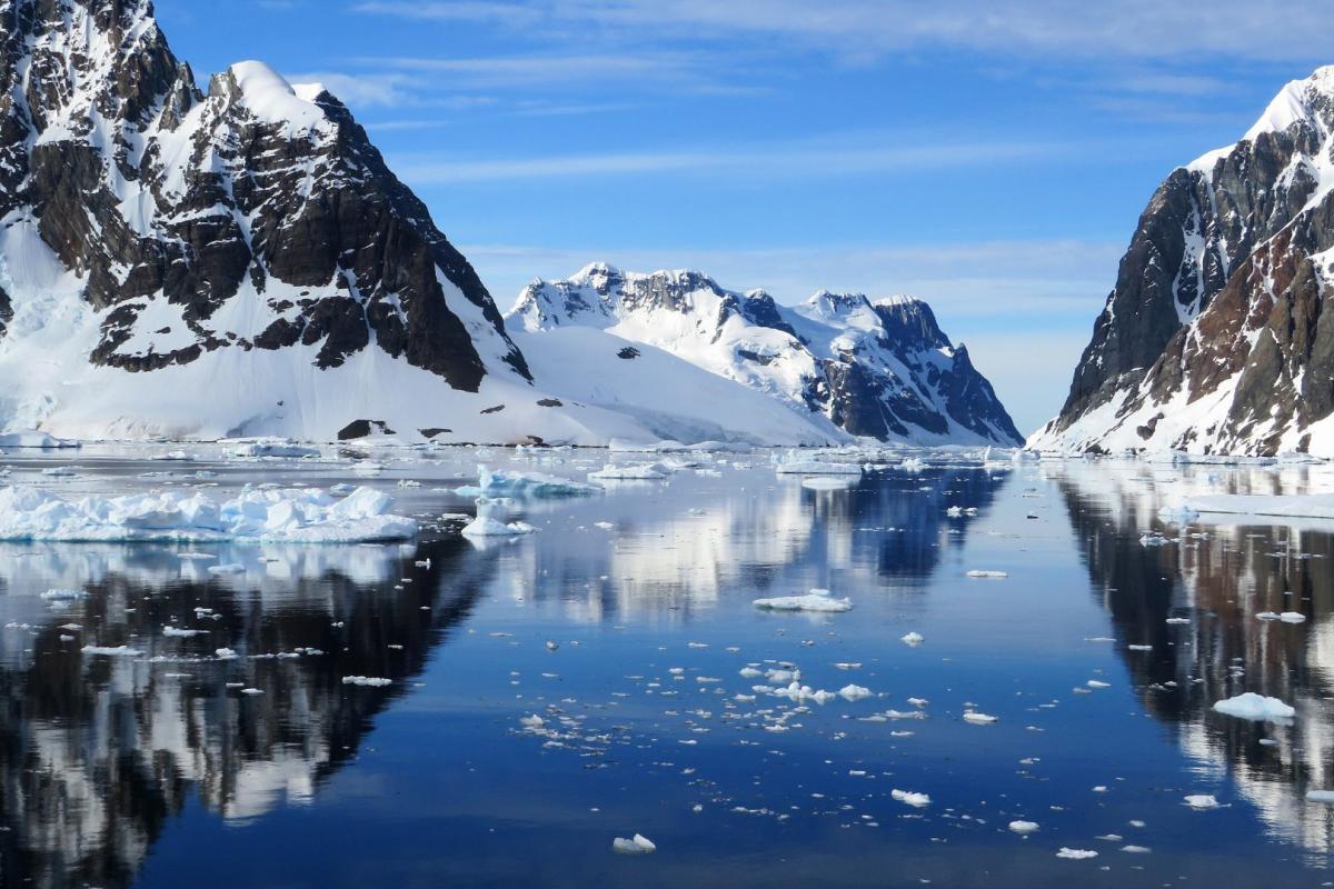 A serene icy landscape shows snow-covered mountains reflecting in the calm waters of a narrow waterway, with floating ice chunks scattered throughout.
