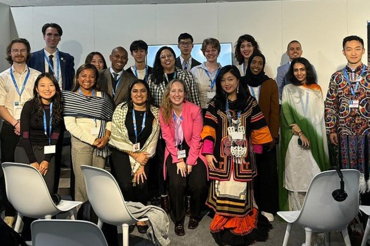 A diverse group of people stand together, some wearing cultural attire, in a conference room with chairs and a banner in the background.