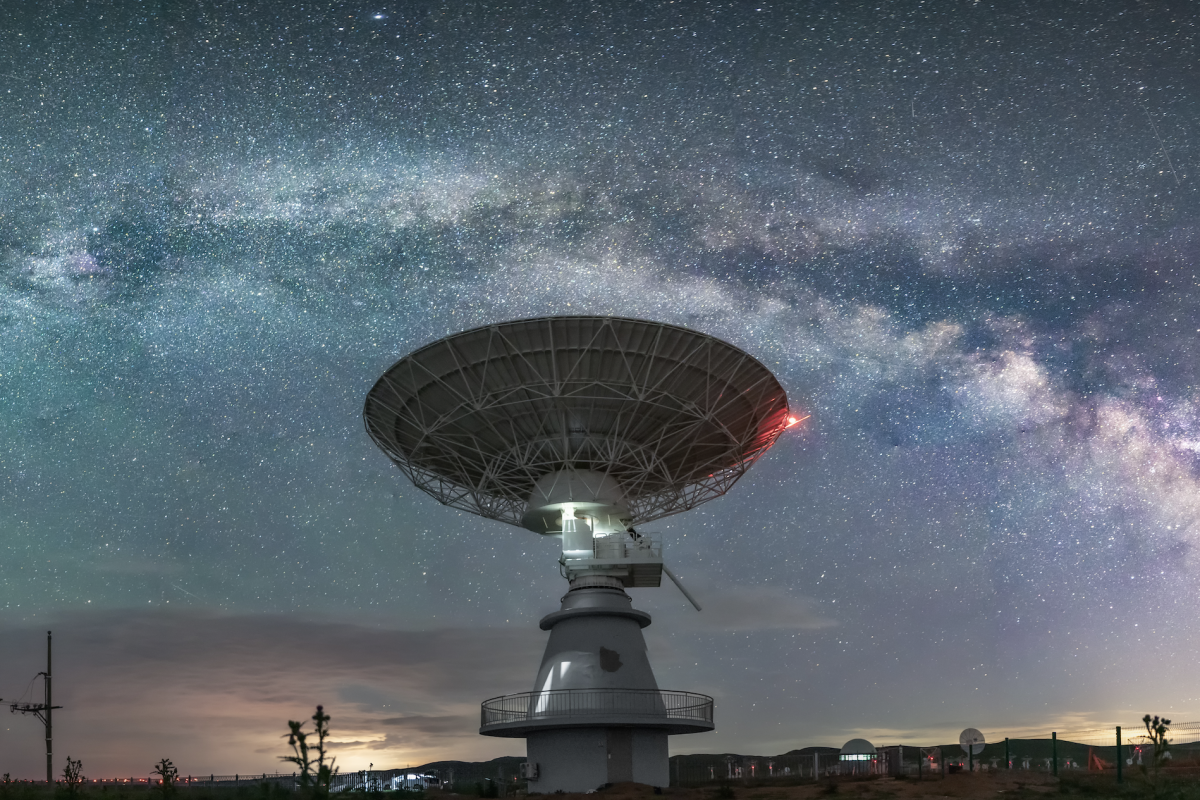 A large radio telescope dish points skyward beneath a clear night sky filled with stars and the arc of the Milky Way galaxy.