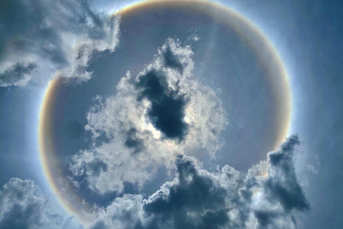 A circular halo surrounds the sun in a cloudy sky, with dark clouds partially obscuring the bright disk.