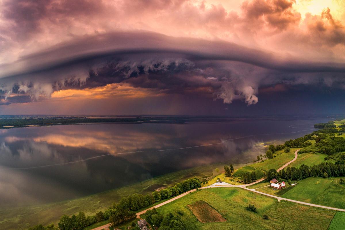 A dramatic storm cloud formation rolls over a calm lake, with green fields and houses in the foreground under a sunset sky.