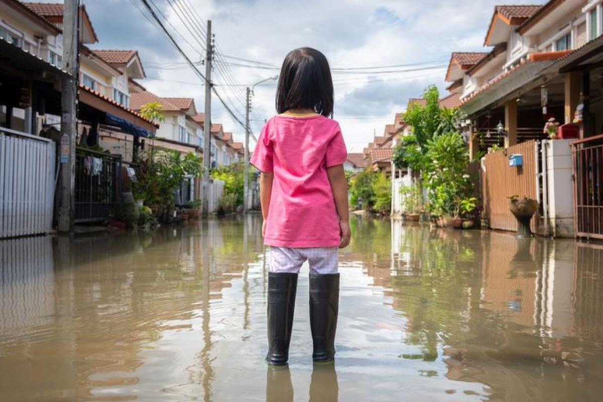 A child in rain boots stands on a flooded street in a residential neighborhood, facing away from the camera.