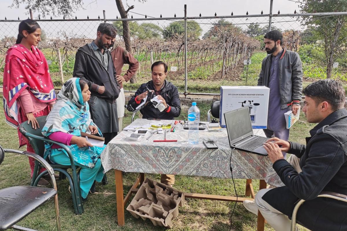A group of people gather around a table outdoors, examining equipment and using a laptop, with demonstration materials and a poster on the table.