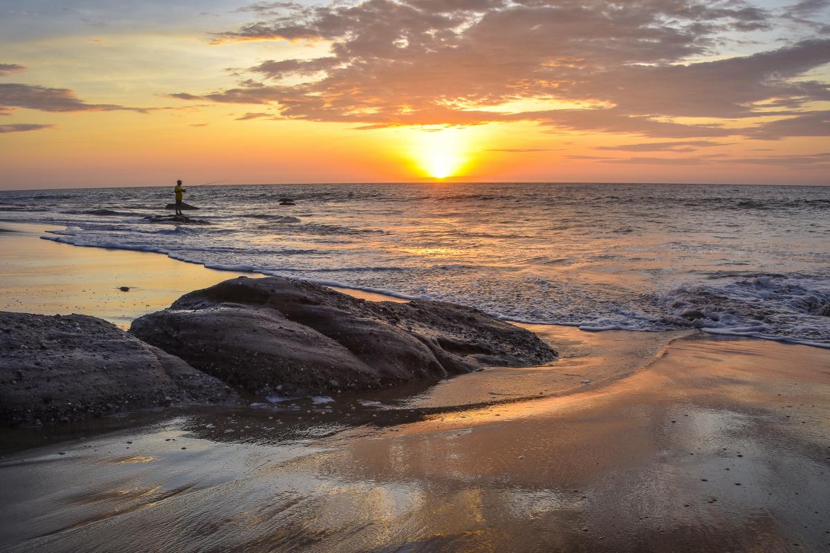 A person on a paddleboard in the ocean at sunset, with rocks and wet sand in the foreground and clouds in the sky.