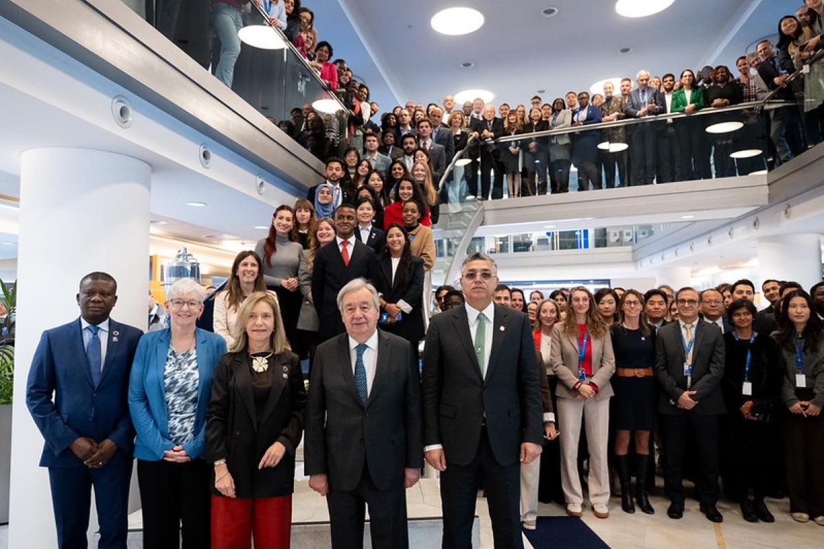 A large group of people, including several formally dressed individuals in front, pose together in a modern, multi-level indoor setting.