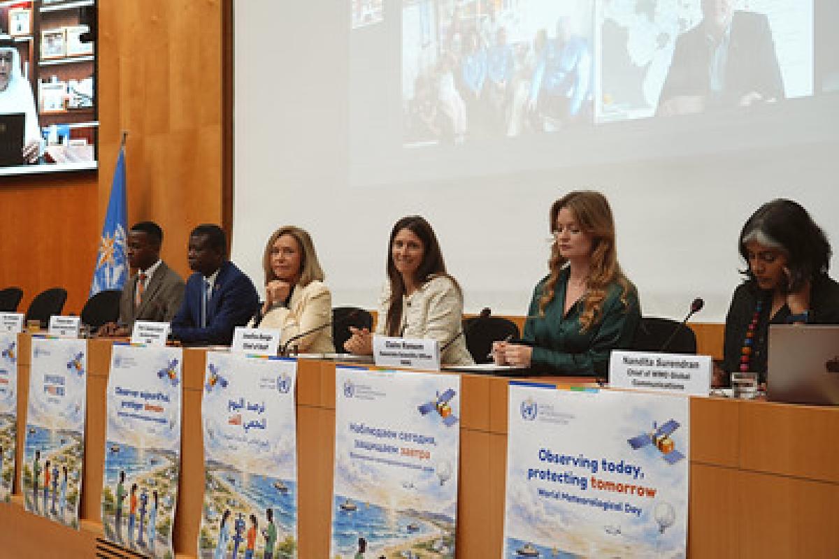Panel of six people seated at a conference table with microphones and nameplates, in front of posters for World Meteorological Day; a video call is visible on a screen behind them.