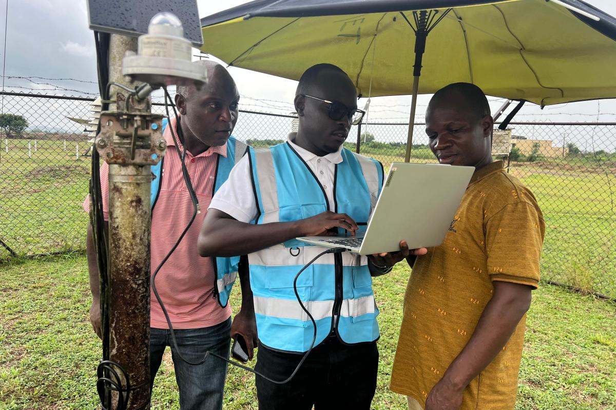 Three men stand outdoors by equipment; one in a blue safety vest uses a laptop, while the others observe. They are under a large umbrella near a fenced grassy area.