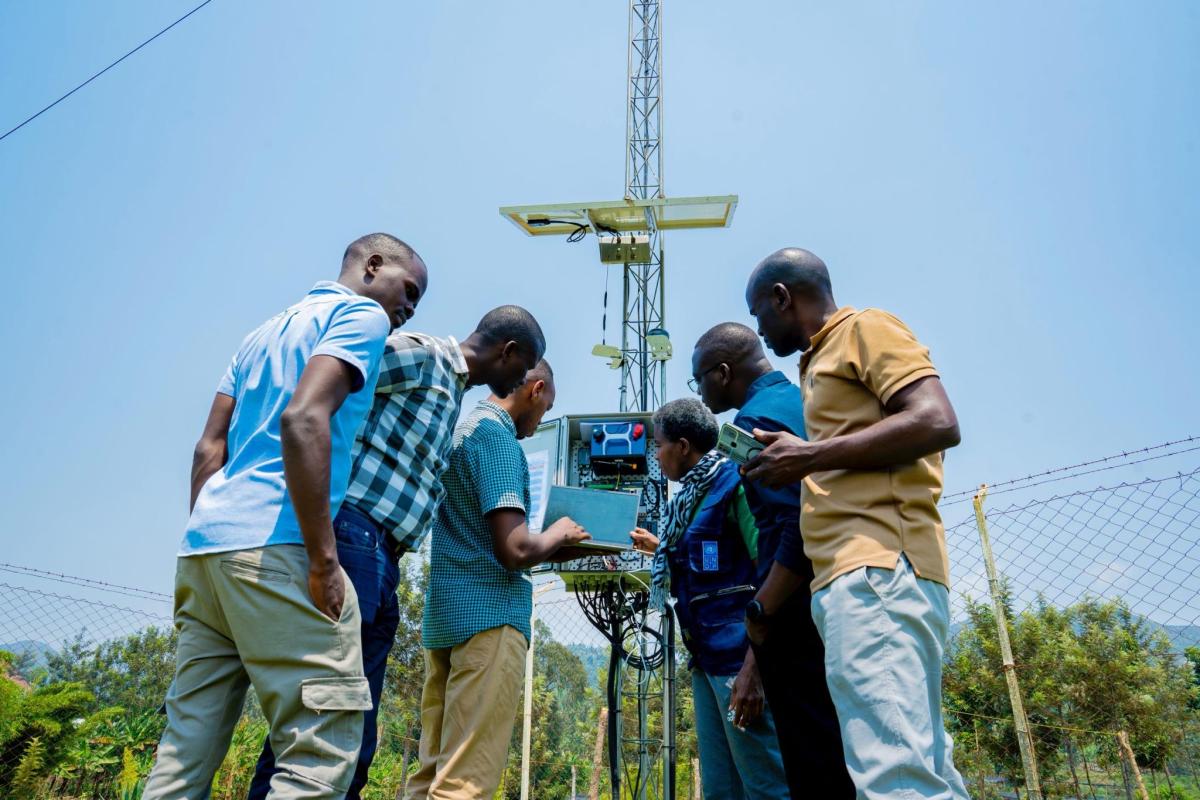 Six people examine and operate a weather monitoring station outdoors near a fence and trees under a clear blue sky.