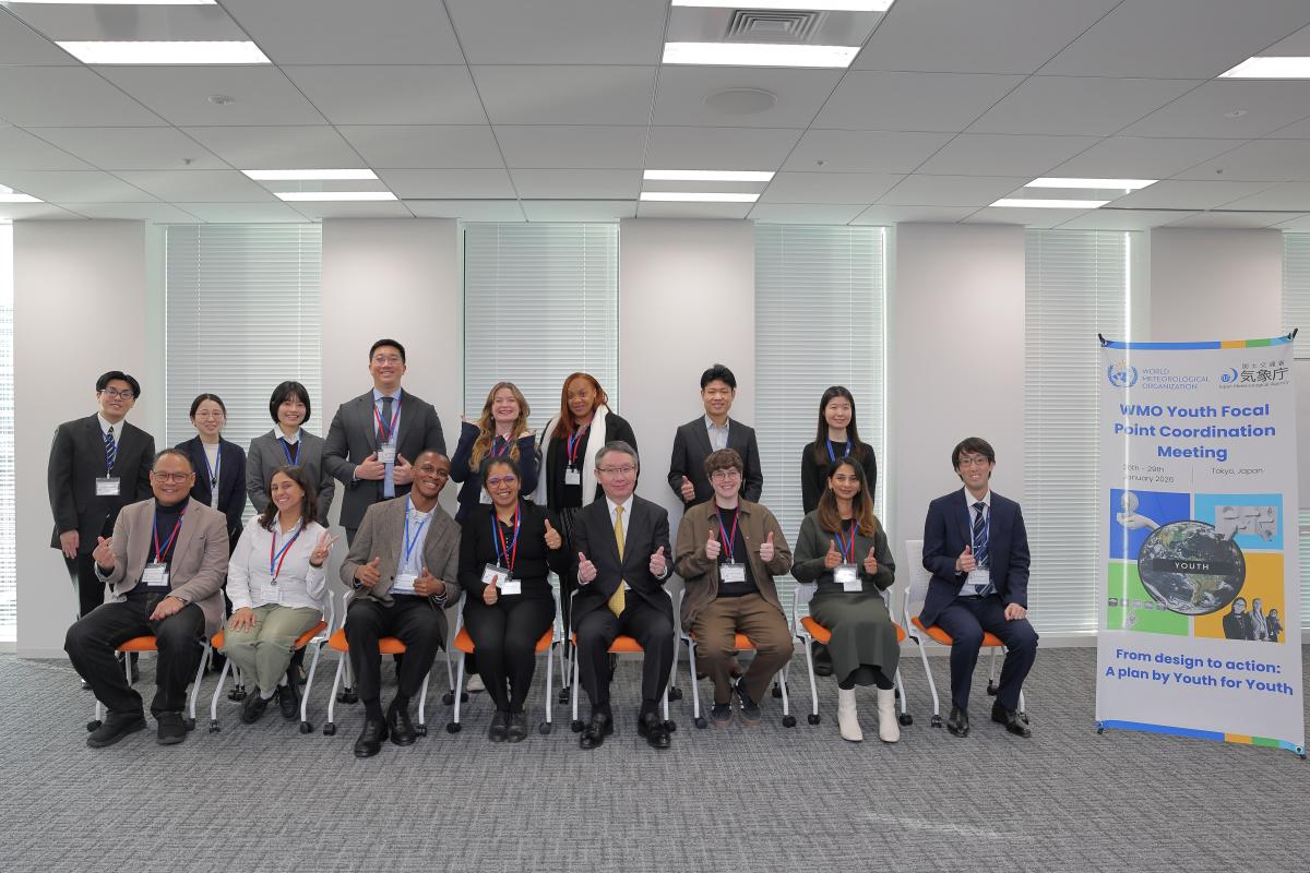 A group of 15 people in business attire pose for a photo in an office setting, seated and standing, next to a banner for a "WMO Youth Focal Point Coordination Meeting.