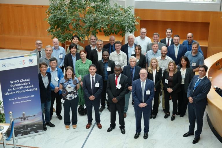 A group of people poses for a photo at the WMO Global Stakeholders on Aircraft-Based Observations Meeting, held indoors with a banner visible in the background.