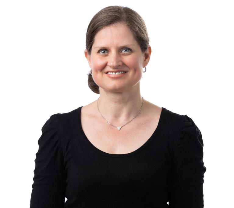 A woman with light skin and brown hair pulled back, wearing a black top, a necklace, and small hoop earrings, smiles at the camera against a white background.