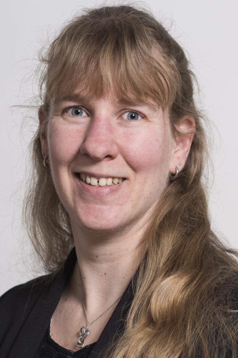 A woman with long, light brown hair and blue eyes, wearing a black top and necklace, smiles at the camera against a plain background.