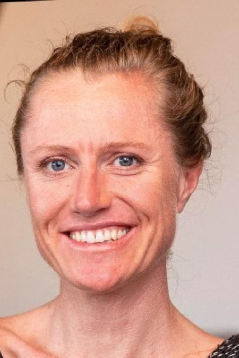 A woman with light skin and blue eyes smiles at the camera. She has wavy blonde hair pulled back and is wearing a patterned top. The background is plain and neutral.