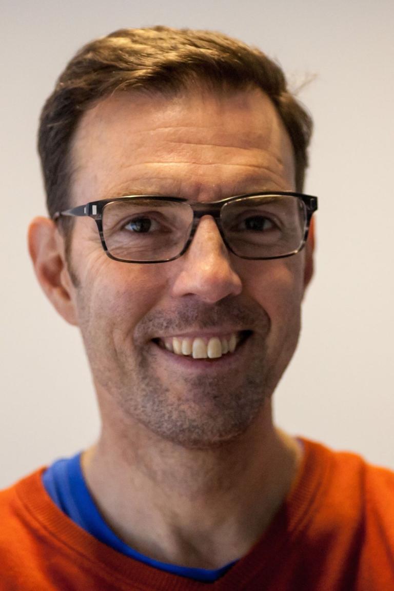 A man with short brown hair, glasses, and a trimmed beard smiles at the camera. He is wearing an orange top over a blue shirt. The background is plain and out of focus.