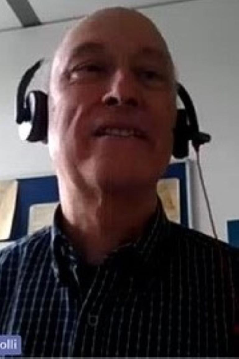 A man wearing a headset and checkered shirt is speaking, possibly during a video call, with documents pinned on the wall behind him.