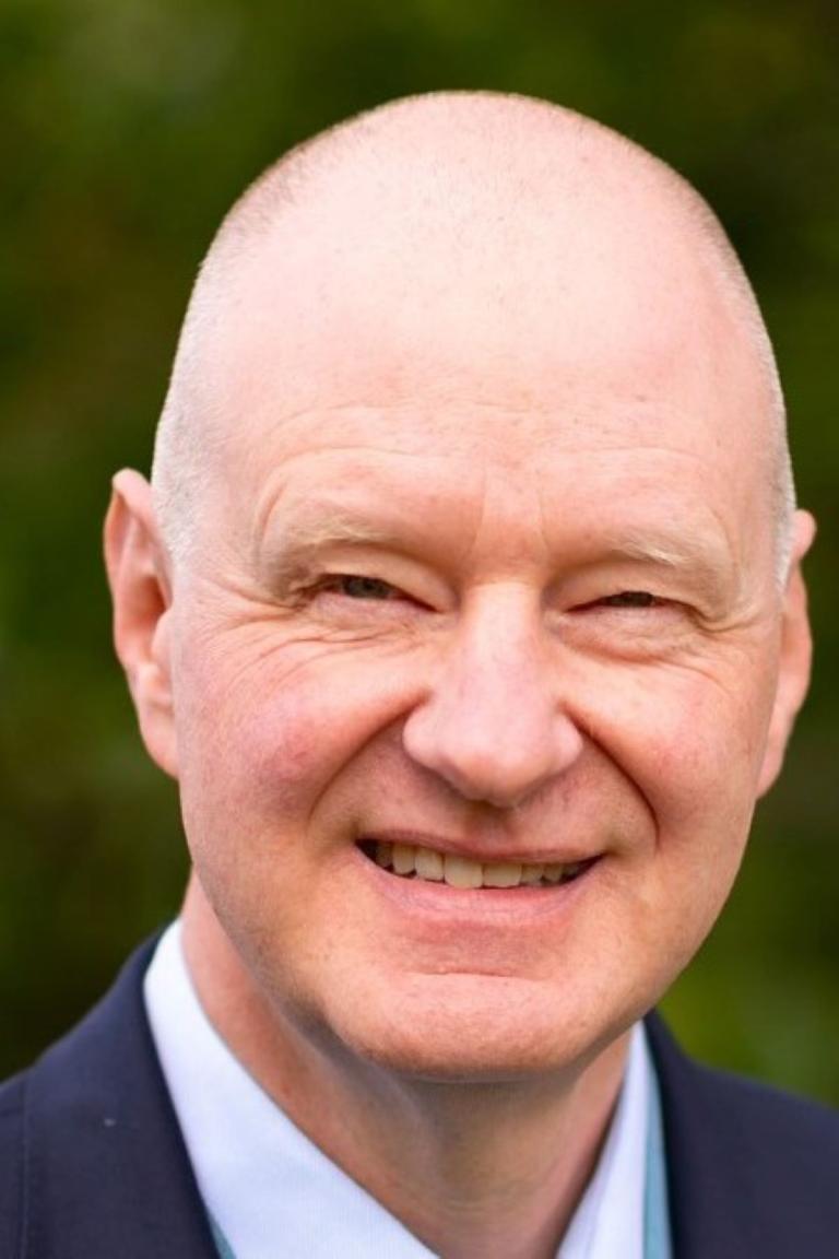 A middle-aged bald man in a suit smiles at the camera with a blurred green background.