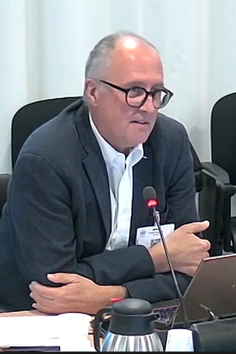 A man wearing glasses and a suit speaks into a microphone at a conference table, with a laptop and a coffee carafe in front of him.