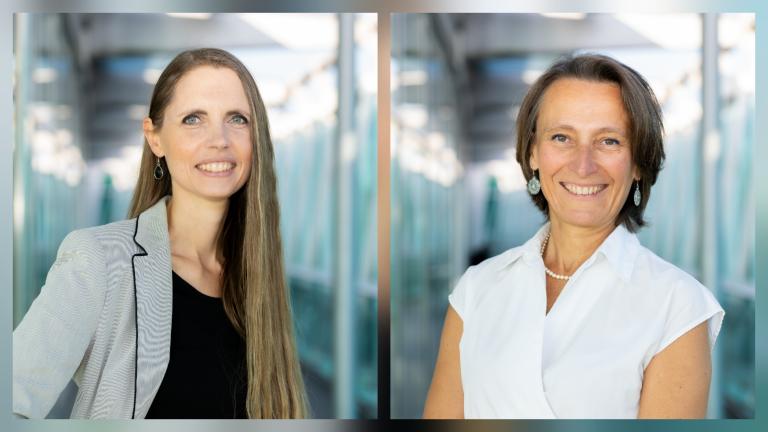 Two women stand in a bright hallway, each posing for a professional portrait. One wears a striped blazer and black top, the other a white blouse. Both are smiling at the camera.
