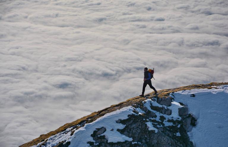 A person with a backpack hikes along a snow-covered mountain ridge above a blanket of clouds.